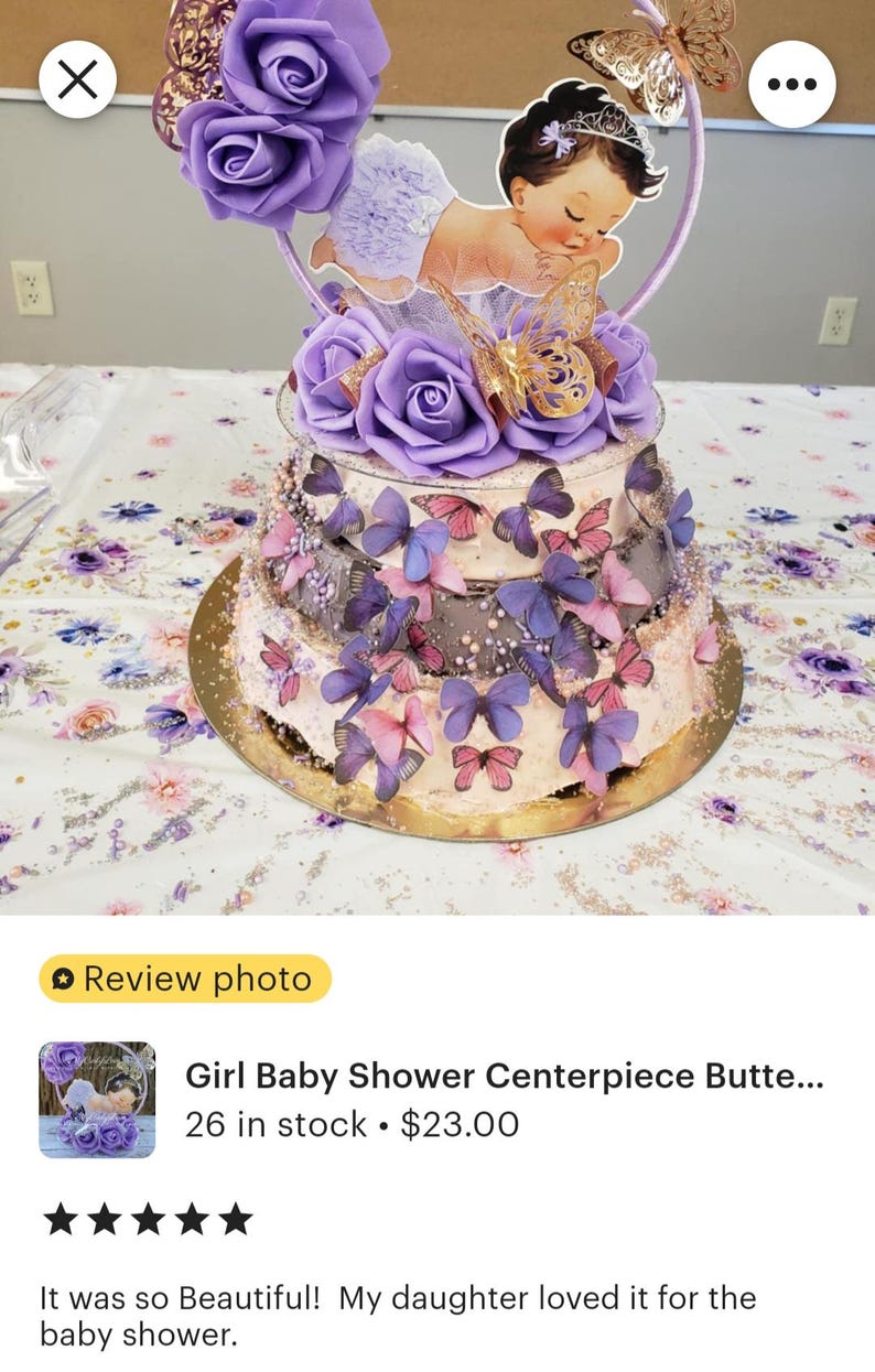 Pode incluir: Um centro de mesa decorativo de v&aacute;rios n&iacute;veis com rosas de espuma roxas, recortes de borboletas com glitter dourado e um recorte central de um beb&eacute; com uma tiara. A estrutura &eacute; adornada com numerosas pequenas borboletas roxas e cor-de-rosa.