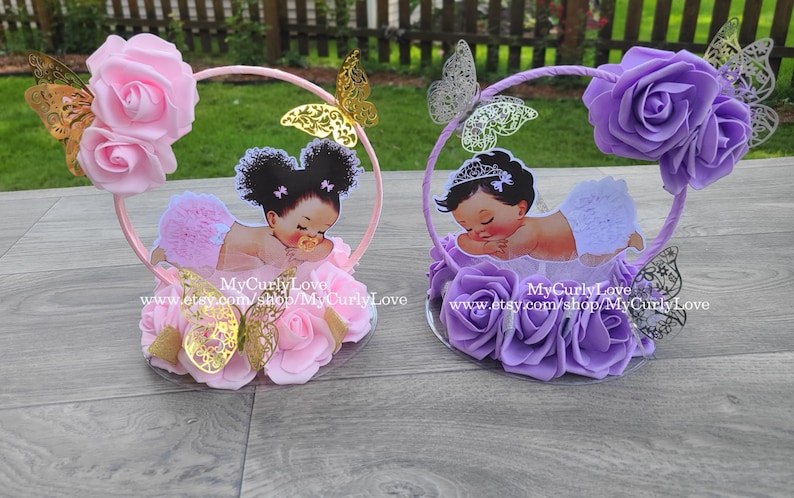Pode incluir: Dois centros de mesa circulares com rosas de espuma, recortes de borboletas douradas e prateadas e ilustra&ccedil;&otilde;es de beb&eacute;s. O centro esquerdo &eacute; rosa com um beb&eacute; de tutu, o direito &eacute; roxo com um beb&eacute; de tiara. O texto diz MyCurlyLove.