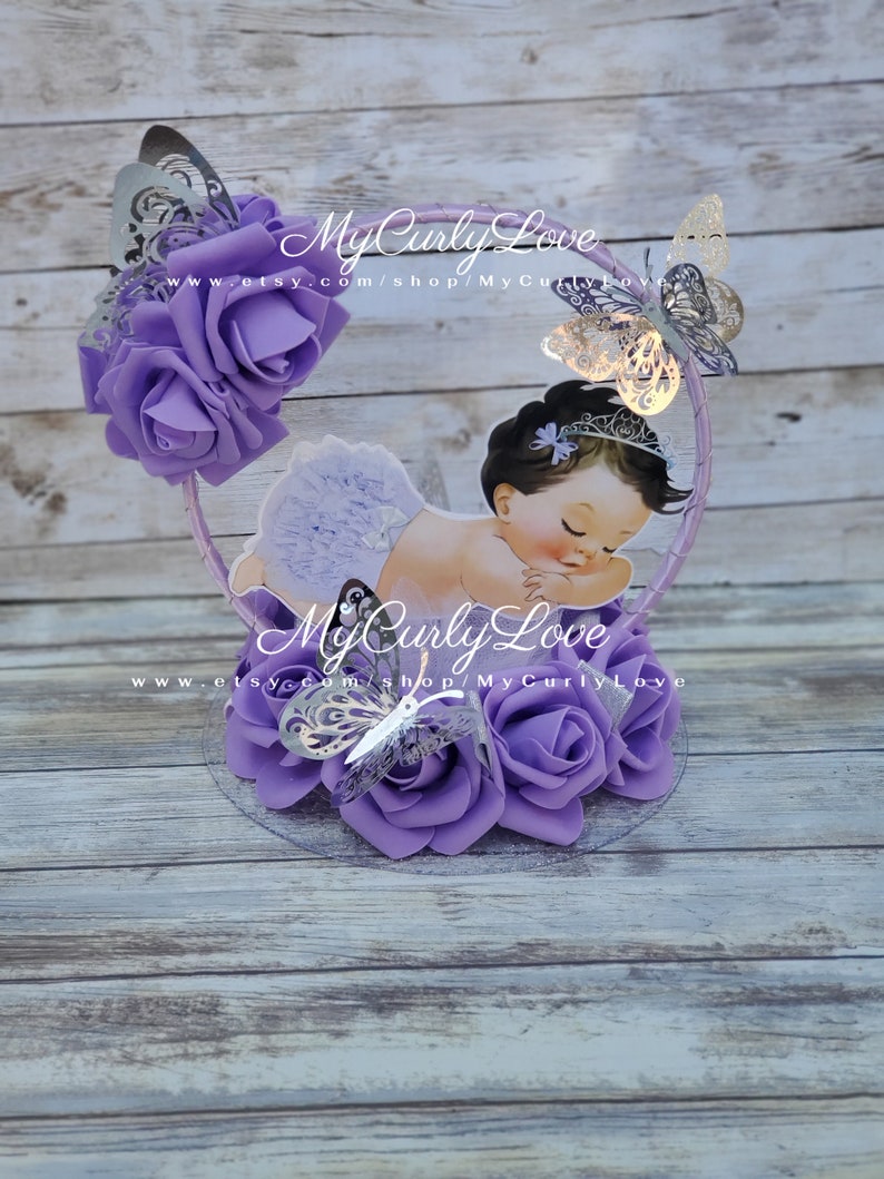 Pode incluir: Um centro de mesa decorativo com a ilustra&ccedil;&atilde;o de um beb&eacute; a dormir com uma tiara e uma saia com folhos, rodeado por rosas de espuma roxas e borboletas prateadas cortadas a laser numa moldura circular com o texto MyCurlyLove impresso.