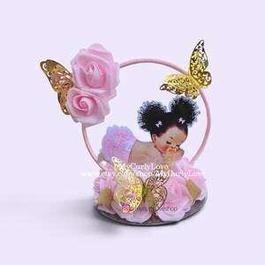 Pode incluir: Um topo de bolo com um aro circular rosa decorado com rosas de espuma rosa claro e recortes de borboletas douradas. Uma figura central mostra um beb&eacute; de cabelo escuro encaracolado com tot&oacute;s, um tutu rosa e uma chupeta.