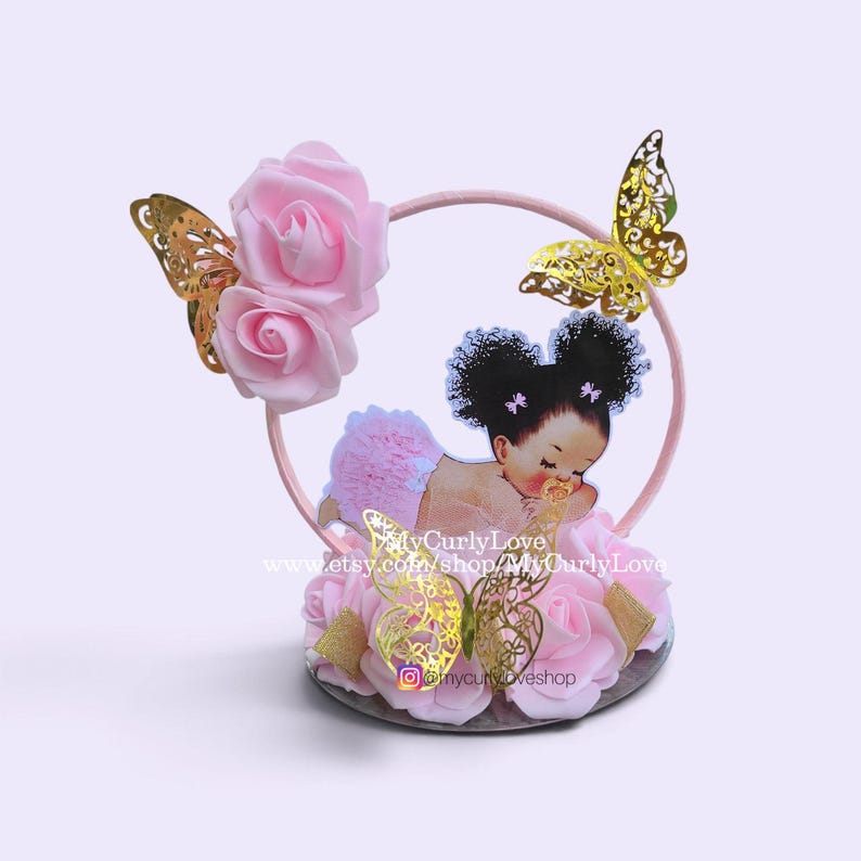 Pode incluir: Um topo de bolo com um aro circular rosa decorado com rosas de espuma rosa claro e recortes de borboletas douradas. Uma figura central mostra um beb&eacute; de cabelo escuro encaracolado com tot&oacute;s, um tutu rosa e uma chupeta.