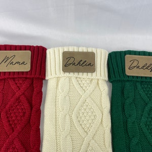 May include: Three knitted stockings with personalized leather tags. The stockings are red, white, and green. The tags read "Mama", "Dahlia", and "Daddy".