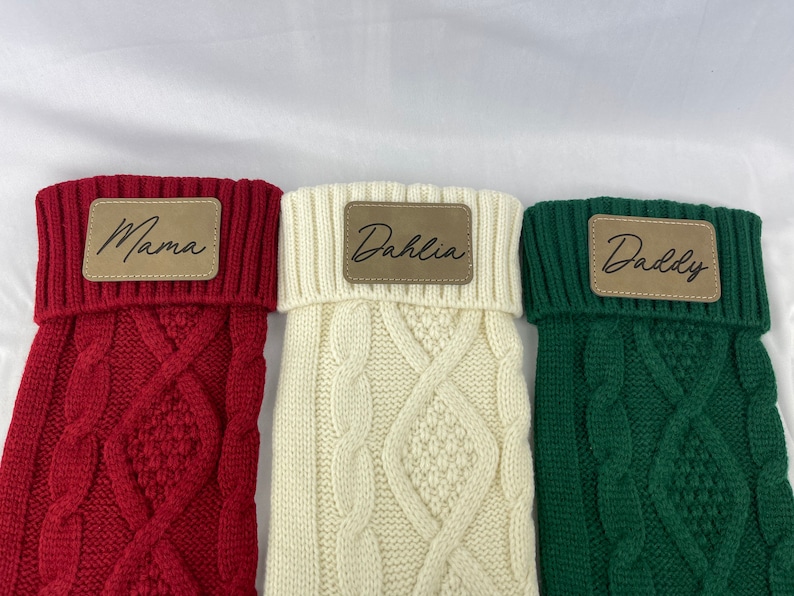 May include: Three knitted stockings with personalized leather tags. The stockings are red, white, and green. The tags read "Mama", "Dahlia", and "Daddy".
