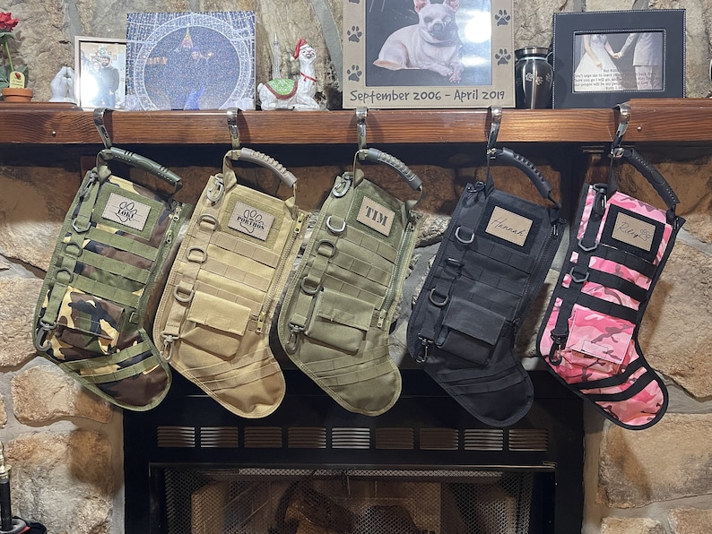 May include: Five personalized camouflage dog stockings hanging on a fireplace mantel. The stockings are in different colors and patterns, including green, tan, black, and pink. Each stocking has a name tag with a paw print.