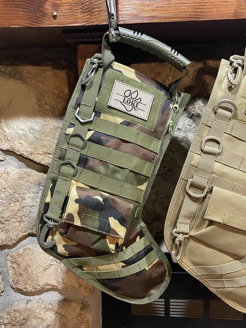 May include: Camouflage-patterned stocking with a black zipper and a green handle. The stocking has multiple straps and a patch with the name "Loki" and a paw print.