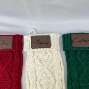 May include: Three personalized knitted Christmas stockings with brown leather name tags. The stockings are red, white, and green. The names on the tags are Eric, Alexis, and Colton.