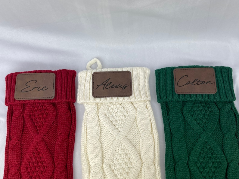 May include: Three personalized knitted Christmas stockings with brown leather name tags. The stockings are red, white, and green. The names on the tags are Eric, Alexis, and Colton.