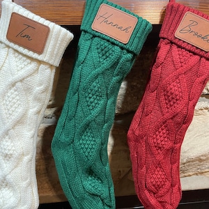 May include: Three knitted Christmas stockings with personalized leather name tags. The stockings are white, green, and red. The names on the tags are Tim, Hannah, and Brooke.