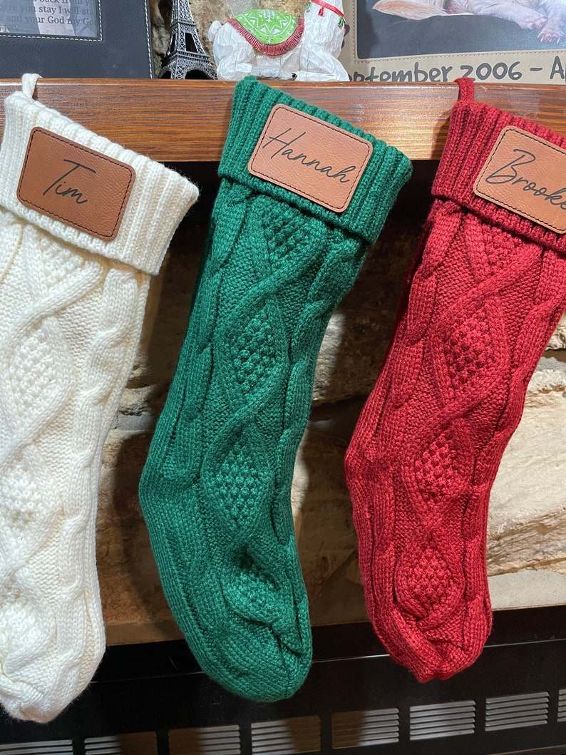 May include: Three knitted Christmas stockings with personalized leather name tags. The stockings are white, green, and red. The names on the tags are Tim, Hannah, and Brooke.