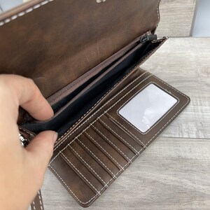May include: A brown leather wallet with a zippered pocket and multiple card slots. The wallet has a white stitching detail and a clear plastic window for displaying an ID card.