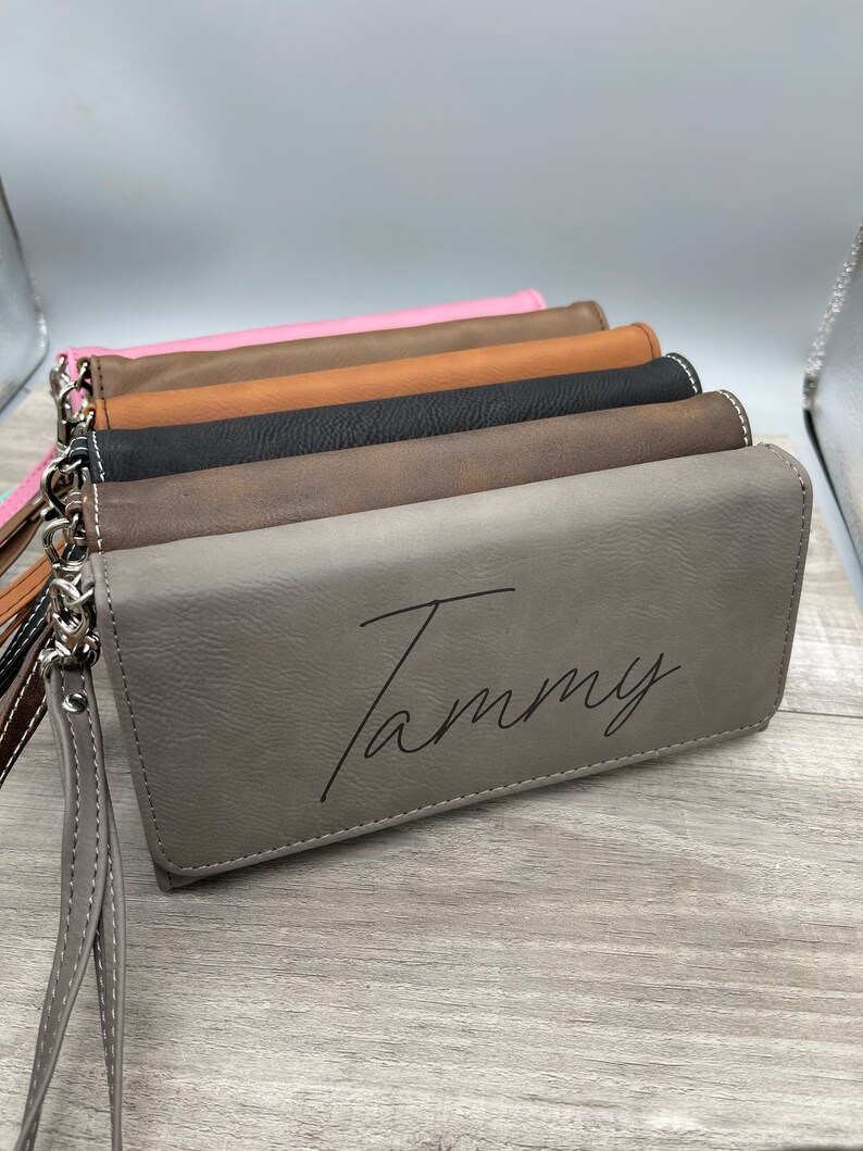 May include: A stack of five different colored leather wallets with a personalized inscription that reads "Tammy" on the gray wallet.