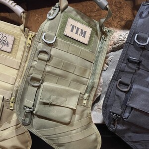 May include: Two green and black tactical style Christmas stockings with a zipper and a handle. The stocking on the left has the name "ETHOS" on it. The stocking on the right has the name "TIM" on it.