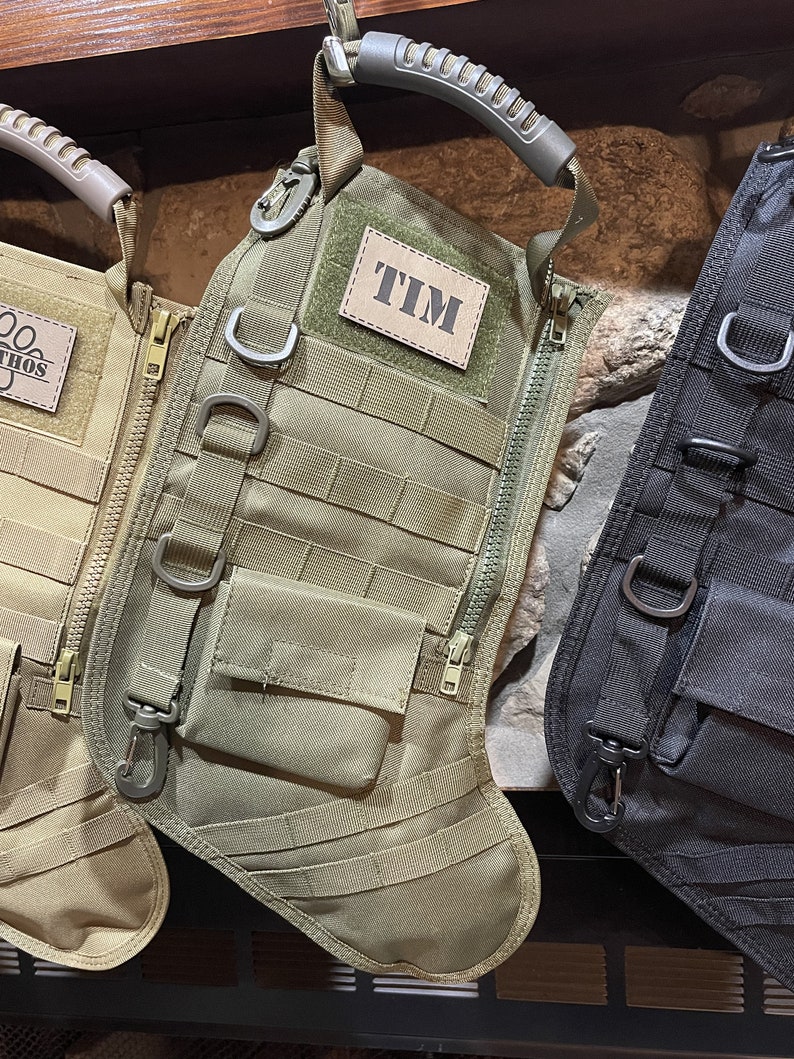May include: Two green and black tactical style Christmas stockings with a zipper and a handle. The stocking on the left has the name "ETHOS" on it. The stocking on the right has the name "TIM" on it.