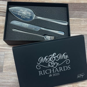 May include: A black gift box with a silver inscription that reads "Mr. & Mrs. Richards Est. 2023" and the words "Little Things" in a smaller font. The box is open, revealing a silver cake knife and server set with the same inscription.