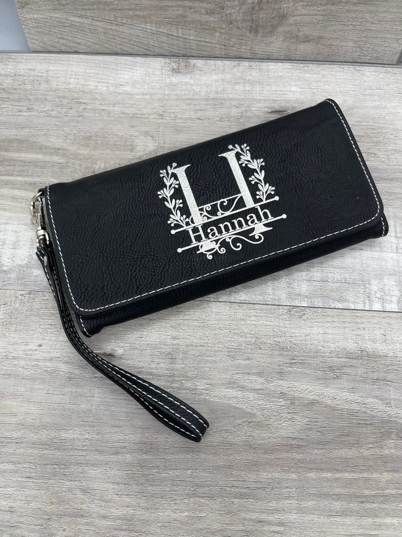 May include: Black faux leather wallet with a white monogram design featuring the letter "H" and the name "Hannah". The wallet has a wrist strap.