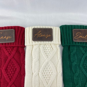 May include: Three knitted stockings with personalized leather name tags. The stockings are red, white, and green. The names on the tags are Caasi, Alonzo, and Emilio.