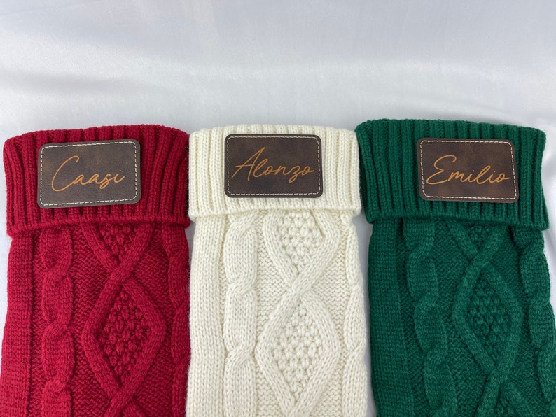 May include: Three knitted stockings with personalized leather name tags. The stockings are red, white, and green. The names on the tags are Caasi, Alonzo, and Emilio.