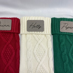 May include: Three knitted stockings with personalized leather name tags. The stockings are red, white, and green. The names on the tags are Quinn, Holly, and Pepere.