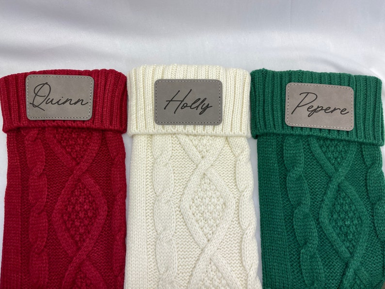 May include: Three knitted stockings with personalized leather name tags. The stockings are red, white, and green. The names on the tags are Quinn, Holly, and Pepere.