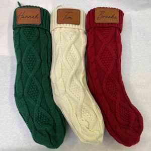 May include: Three Christmas stockings with personalized leather name tags. The stockings are made of a thick, cable knit fabric. The stockings are green, white, and red.