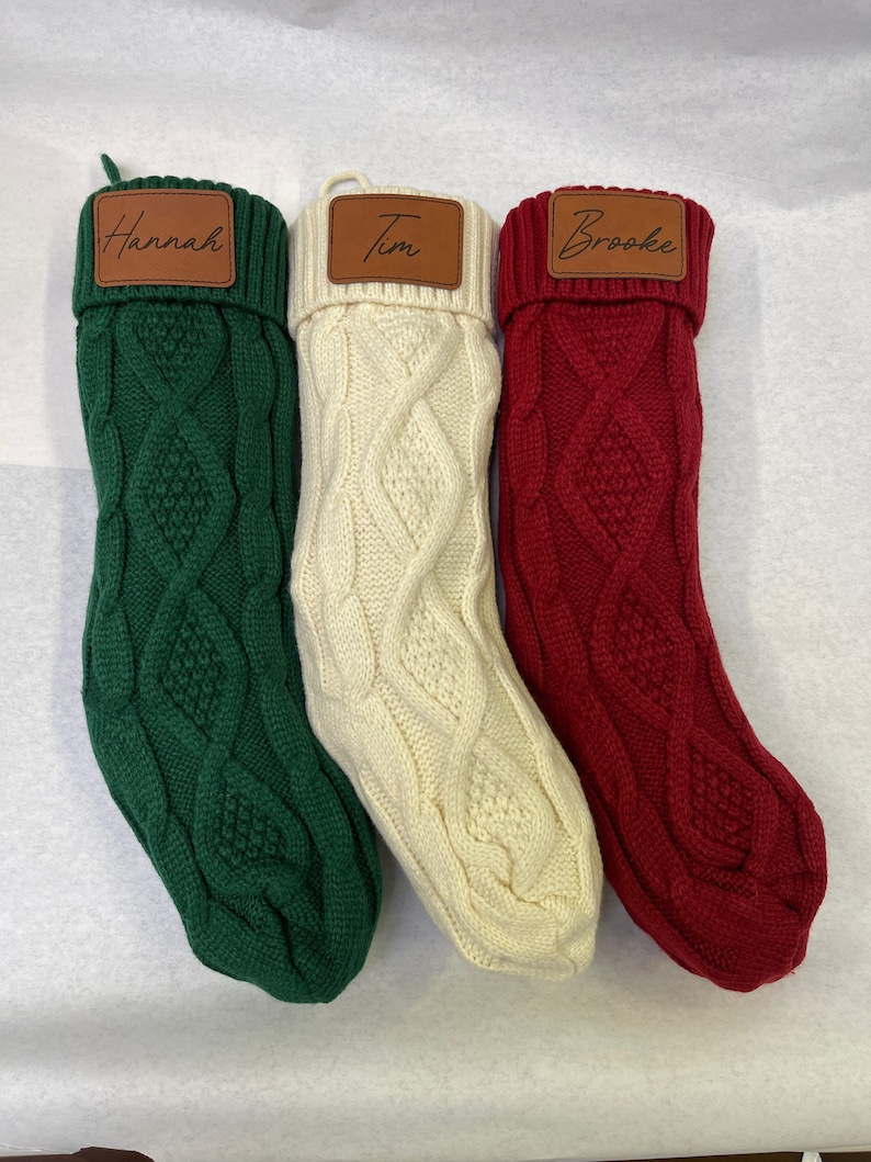 May include: Three Christmas stockings with personalized leather name tags. The stockings are made of a thick, cable knit fabric. The stockings are green, white, and red.