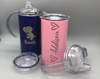 Puede incluir: Dos vasos de acero inoxidable con tapas y asas. El vaso azul tiene un gráfico de dinosaurio y el texto "Doodle". El vaso rosa tiene el texto "Addison" con un corazón.