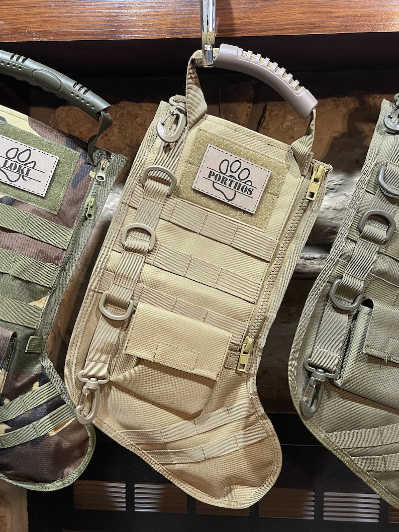 May include: Tan-colored, tactical-style dog stocking with a zipper, multiple pockets, and a loop handle. The stocking has a personalized patch with the name "PORTHOS" and a paw print.
