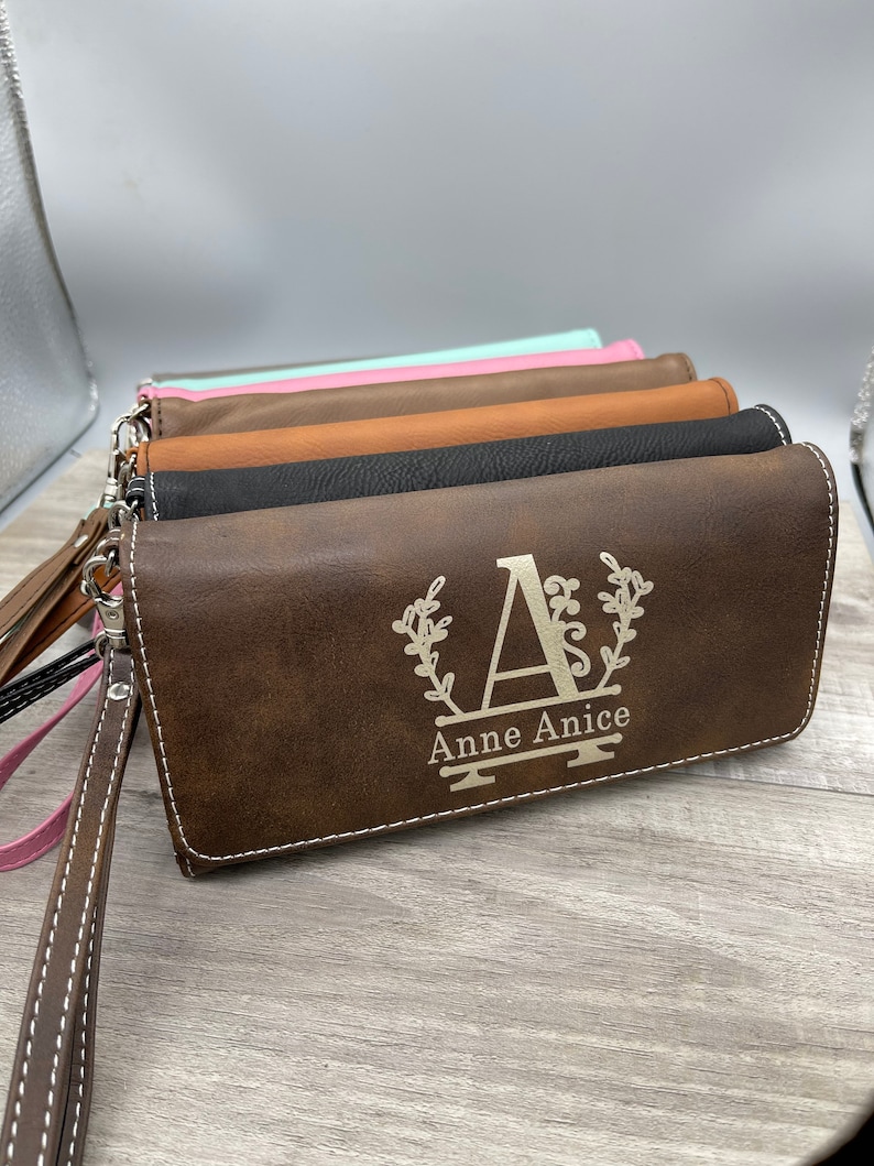 May include: A stack of brown leather wallets with a personalized monogram. The top wallet has the letter "A" with a floral design and the name "Anne Anice" engraved on it.