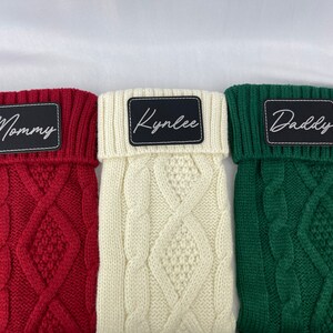 May include: Three knitted dog sweaters with personalized names. The sweaters are red, white, and green with a cable knit pattern. The names are "Mommy", "Kynlee", and "Daddy".