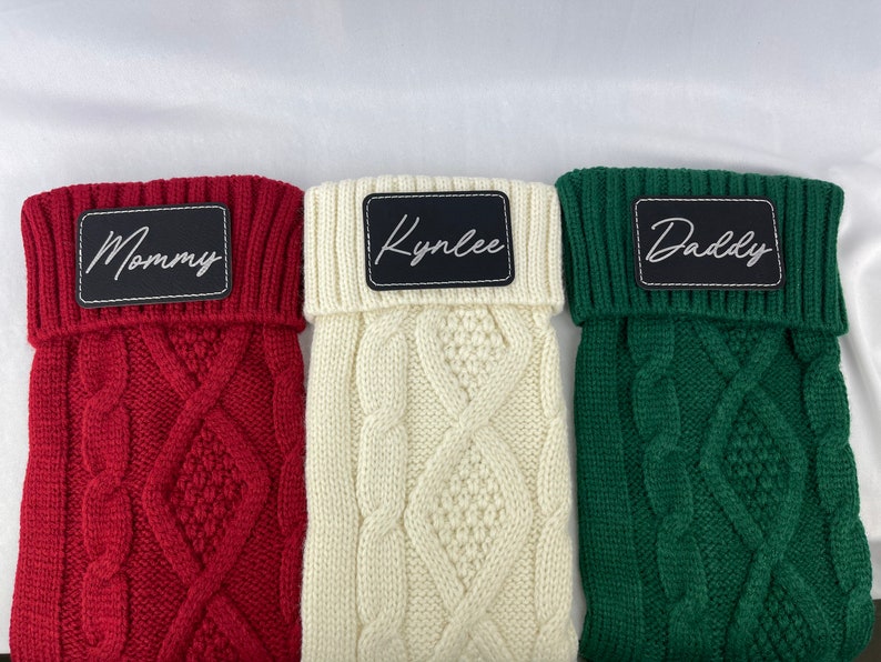 May include: Three knitted dog sweaters with personalized names. The sweaters are red, white, and green with a cable knit pattern. The names are "Mommy", "Kynlee", and "Daddy".