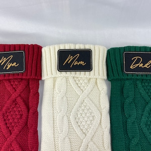 May include: Three knitted Christmas stockings with personalized names. The stockings are red, white, and green. The names are "Mya", "Mom", and "Dad" in gold lettering.