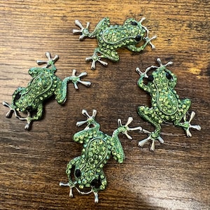May include: Four green jeweled frog brooches with black eyes. The frogs are made of metal and have a shiny finish.