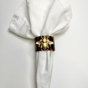 May include: A brown and gold napkin ring with a bee design. The ring is made of resin and features a gold-toned bee with wings.