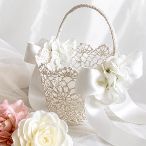 May include: A white, crocheted flower girl basket with a handle and white satin ribbon bows. The basket is decorated with white flowers and is set against a soft, white background. The basket is approximately 8 inches tall.
