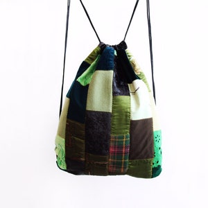 May include: A patchwork drawstring backpack in a green, brown and blue colour scheme. The bag is made from various fabrics and has a unique, handmade appearance.