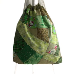 Op de afbeelding: Groene patchwork trekkoordtas met een Hello Kitty-ontwerp. De tas is gemaakt van verschillende groene stoffen met verschillende patronen.