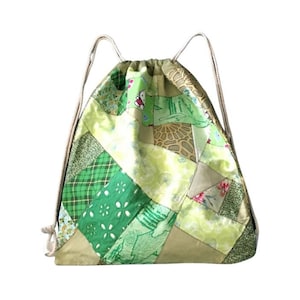 Op de afbeelding: Een groene en beige patchwork trekkoordtas met een wit koord. De tas is gemaakt van verschillende stoffen met verschillende groene en beige patronen.