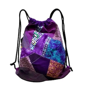 Op de afbeelding: Paarse trekkoordtas gemaakt van patchwork stof met verschillende patronen en texturen. De tas heeft een zwart trekkoord en is perfect voor het dragen van kleine items.