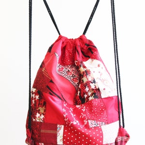 May include: Red patchwork drawstring backpack with black drawstrings. The bag is made of different red fabrics with various patterns.