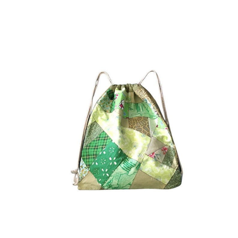 Op de afbeelding: Een groene en beige patchwork trekkoordtas met een wit koord. De tas is gemaakt van verschillende stoffen met verschillende groene en beige patronen.