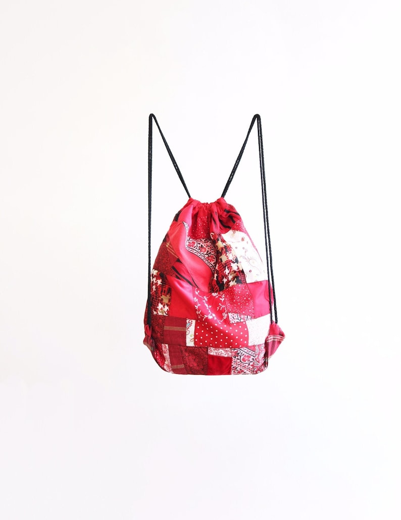 May include: Red drawstring backpack with a patchwork design. The bag is made of fabric with a variety of red patterns.