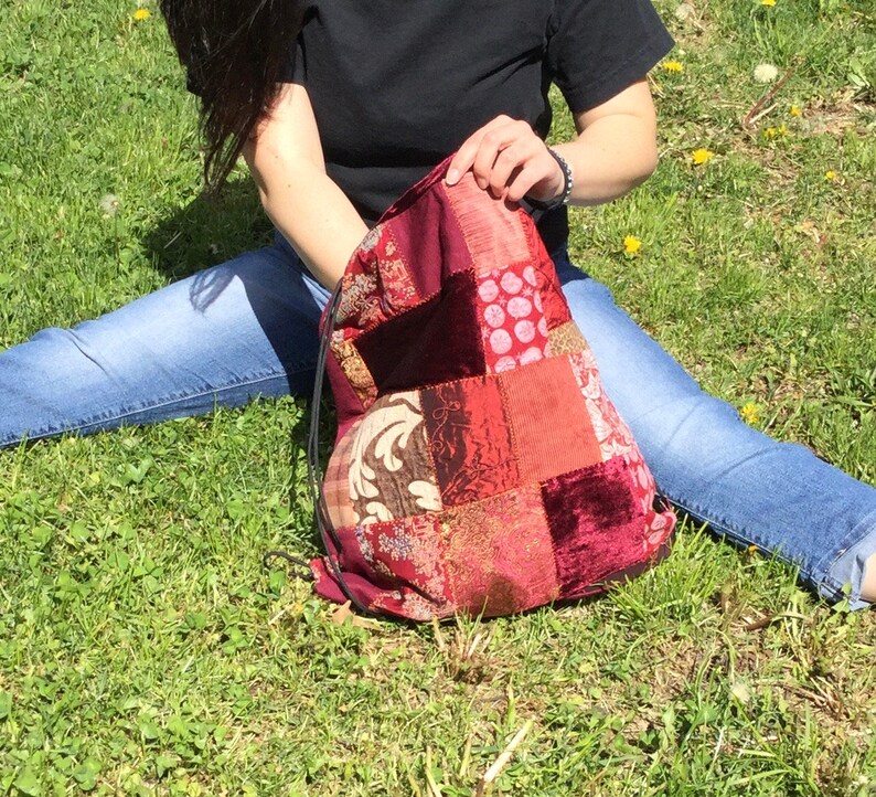 Op de afbeelding: Een rode, bruine en witte patchwork trekkoordtas met een fluwelen textuur. De tas is gemaakt van verschillende stoffen en heeft een unieke, bohemian stijl.