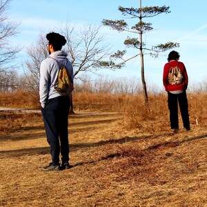 May include: Two people wearing backpacks stand in a field with dry grass and trees in the background. One person is wearing a gray sweatshirt and the other is wearing a red sweatshirt.