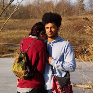 May include: A couple standing close together outdoors. The person on the left is wearing a red sweater and a green and brown patterned backpack. The person on the right is wearing a light blue hoodie and a red and black patterned bag.