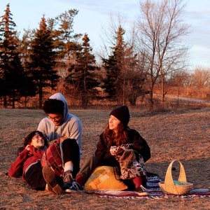 May include: Three people are sitting on a grassy field with a picnic basket. Two people are wearing hooded sweatshirts and one person is wearing a black beanie. The person in the beanie is holding a cup. The people are sitting on a blanket with pillows.