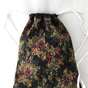 Puede incluir: Una bolsa de cord&oacute;n negro con un patr&oacute;n de tapicer&iacute;a floral en tonos rojos, verdes y dorados. La bolsa tiene cordones de color blanco.