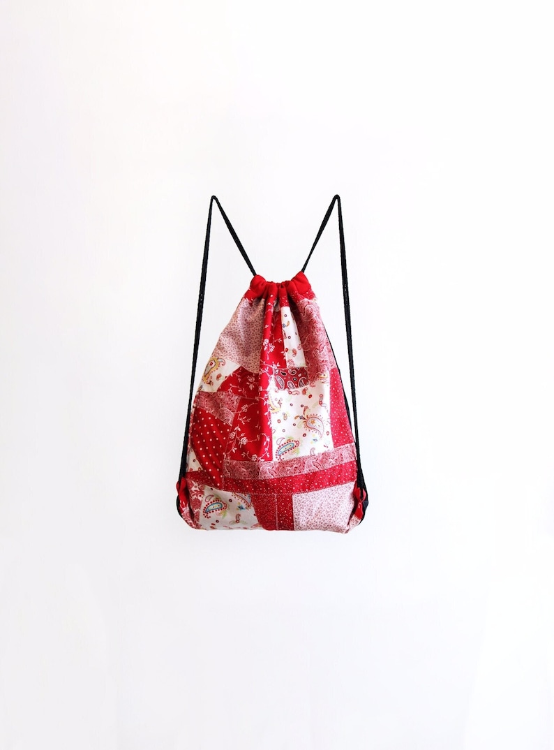 May include: Red and white patchwork drawstring bag with black drawstrings. The bag is made of different fabrics with floral and paisley patterns.
