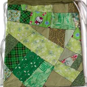 Op de afbeelding: Een groene trekkoordtas gemaakt van patchwork stof. De tas is gemaakt van verschillende groene stoffen met verschillende patronen, waaronder bloemen, ruitjes en geometrische patronen. Op een van de stofvierkanten staat een klein Hello Kitty-figuur.