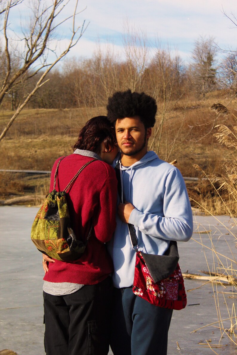 May include: A couple standing close together outdoors. The person on the left is wearing a red sweater and a green and brown patterned backpack. The person on the right is wearing a light blue hoodie and a red and black patterned bag.