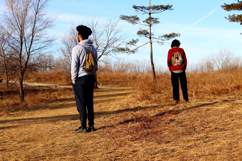 May include: Two people wearing backpacks stand in a field with dry grass and trees in the background. One person is wearing a gray sweatshirt and the other is wearing a red sweatshirt.